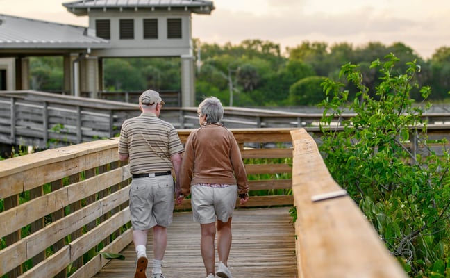 Elderly couple walking hand in hand along a wooden boardwalk through Florida wetlands at sunset