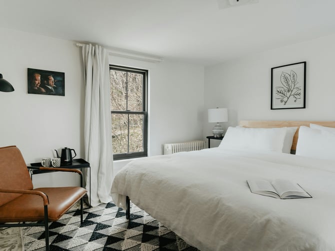 Clean white private bedroom with leather chair, open book on bed, and natural light through a curtained window