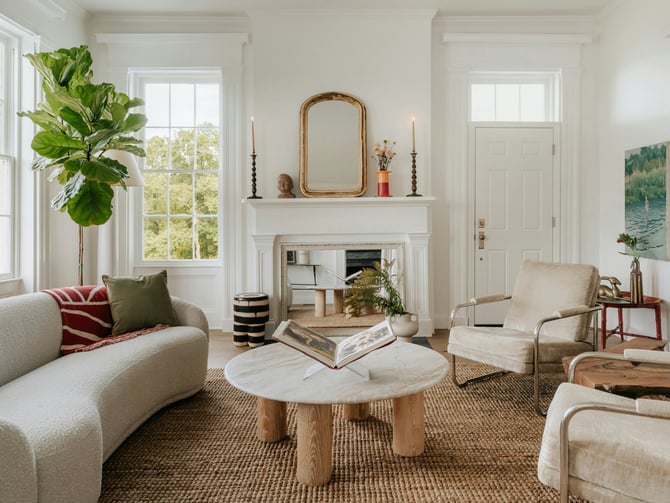 Bright white shared living room with curved sofa, fireplace, fiddle-leaf fig, and large windows overlooking trees